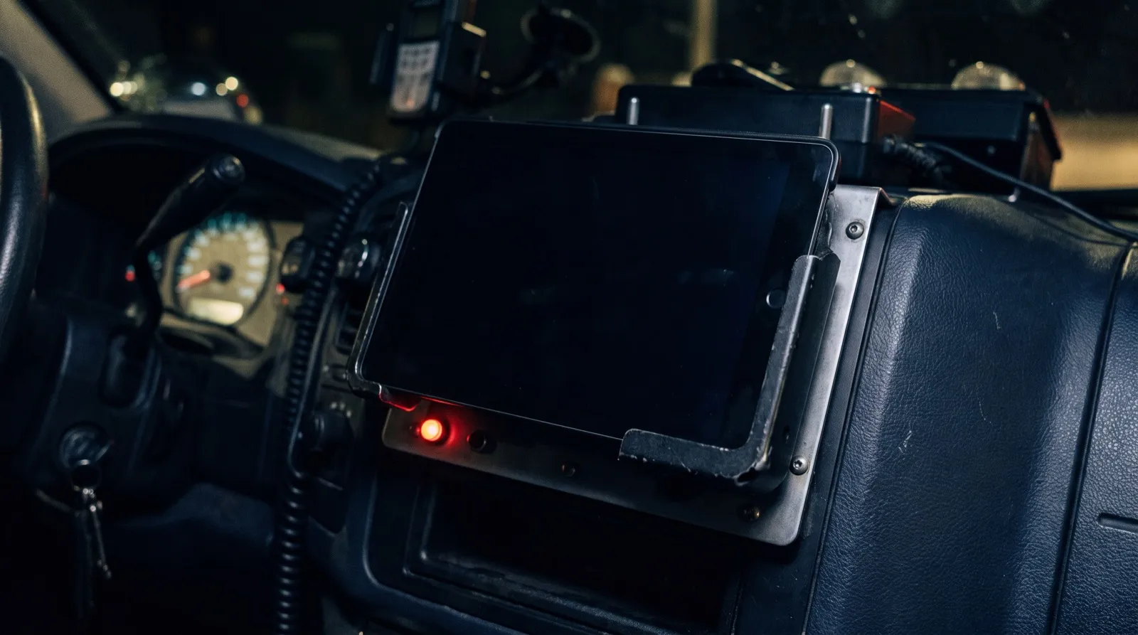 Tablet with a dark screen held in a metal dashboard cradle inside an ambulance cab at night, with a small red power light glowing below it